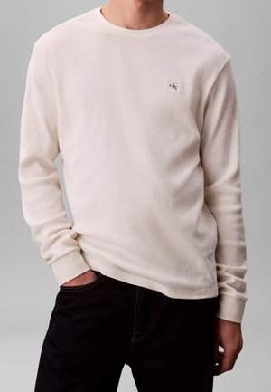 Mann trägt ein cremefarbenes, strukturiertes Langarmhemd mit kleinem Calvin Klein-Logo auf der Brust und dunkle Hose, posiert mit einer Hand in der Tasche.