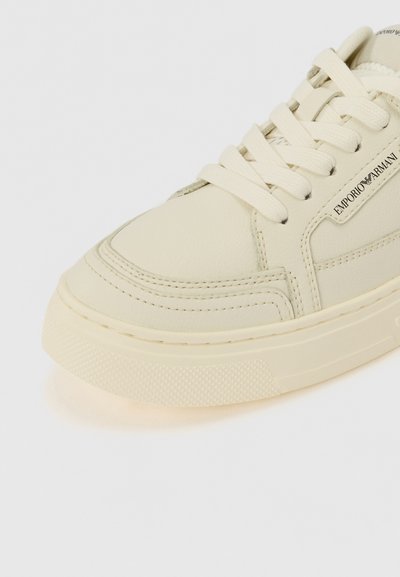 Cream leather sneaker with round toe, textured sole, tonal stitching, and lace closure. Features branded accents on the side.