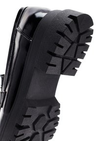 Scarpa in finta pelle nera con suola in gomma testurizzata, caratterizzata da scanalature profonde e tacco piatto. Cuciture laterali visibili vicino alla caviglia.