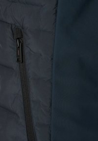 Dunkelblau gesteppte Daunenjacke, links mit einem gesteppte Textur und rechts mit einem glatten Stoff, ausgestattet mit einem seitlichen Reißverschluss und schwarzen Beschlägen.