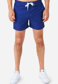 Pantaloni da bagno blu con vita regolabile, design semplice, orlo piegato e due tasche laterali, abbinati a sneakers bianche.