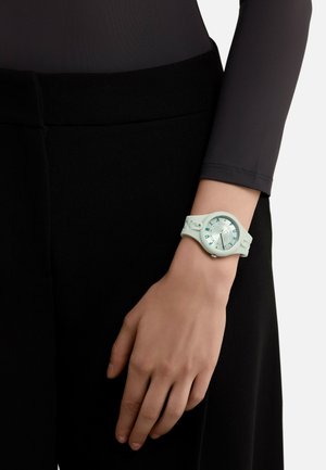 Orologio in silicone verde menta indossato su un polso, con quadrante rotondo di colore blu chiaro, dettagli in argento e indicatori dell'ora ben leggibili.