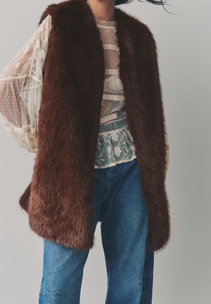 Personne portant un gilet en fourrure marron foncé par-dessus un chemisier en dentelle transparente à motifs floraux et un jean bleu, sur un fond clair uni.