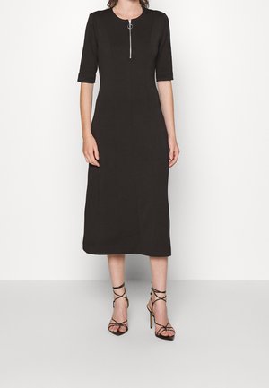 Femme portant une robe midi noire avec des manches aux coudes et une fermeture éclair à l'avant, associée à des sandales noires à talons hauts avec lanières.