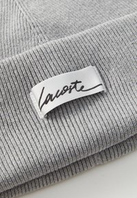 Grå strikket hue med ribbet tekstur, med et hvidt label med sort skrifttype "Lacoste" logo; blødt stof, dobbelt cuff.
