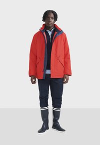 Rote wasserdichte Jacke mit navyfarbenen Akzenten, Kapuze und zwei vorderen Taschen; kombiniert mit navyfarbenen Hosen und hohen navyblauen Gummistiefeln mit weißen Streifen.