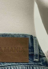 Džínsy s modrým prateľným efektom, s hnedou koženou nášivkou s nápisom "A MANIA." Textúrovaná látka a tradičný dizajn s piatimi vreckami.