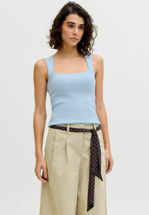 Femme aux cheveux bruns bouclés portant un top sans manches côtelé bleu clair et un pantalon beige taille haute avec une ceinture noire à pois.