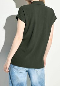 Tmavě zelená košile s krátkým rukávem vyrobená z texturované tkaniny a s límečkem, kombinovaná s světle modrými džínami. Košile jemně splývá z ramen.