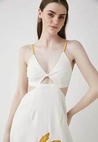 Weißes Kleid mit gelben Trägern, V-Ausschnitt und einem Frontauschnitt. Besitzt ein abstraktes, gelb gesticktes Detail am Saum. Glatte Textur.