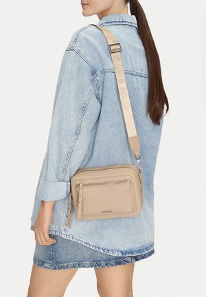 Mujer con chaqueta y falda de mezclilla clara llevando un bolso cruzado beige con una correa ancha y ajustable sobre el hombro.