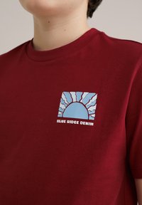 Maroon majica od pamuka s plavim grafičkim prikazom izlazećeg sunca s zracima. Ispod grafike je prikazan tekst "BLUE RIDGE DENIM".