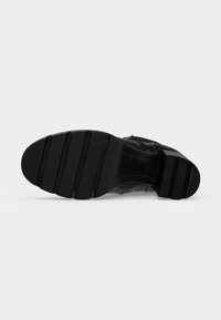 Semelle de chaussure noire avec des rainures horizontales et un talon bas et large, vue du dessous sur un fond clair uni.