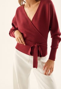 Donna che indossa un maglione avvolto a maniche lunghe color bordeaux a costine, legato in vita, abbinato a pantaloni bianchi, mostrata dalla regione del petto alle cosce.