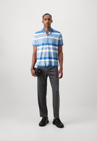 Camicetta a maniche corte a righe blu chiaro e bianche, abbinata a pantaloni grigi scuri su misura e sneakers nere. Il modello tiene una piccola borsa nera.
