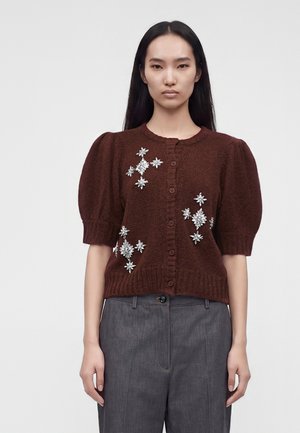 Mujer con un cárdigan marrón de manga corta con adornos florales plateados, pantalones grises de pierna ancha y zapatos con plataforma marrones, de pie con la mano en la cadera.
