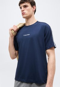 Νεαρός άνδρας που φοράει μπλε μαρινιέ μπλουζάκι Calvin Klein με ριγέ μανίκια, κρατώντας μπεζ σακάκι στους ώμους, μπροστά σε απλό φόντο.