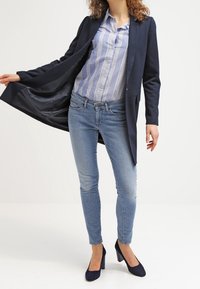 Marineblauer langer Blazer über einem blau-weiß gestreiften Hemd mit Knöpfen, kombiniert mit figurschmeichelnden hellblauen Jeans und marineblauen High Heels. Elegantes, schlankes Design.