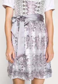 Eine Frau trägt ein silberlila Dirndlkleid mit einer floralem Spitze-Apron und einer breiten Satinschleife, die auf der rechten Seite der Taille gebunden ist.