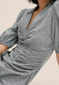 Femme portant une robe grise texturée avec des manches courtes bouffantes et des détails de tissu froncé sur le devant et la taille.