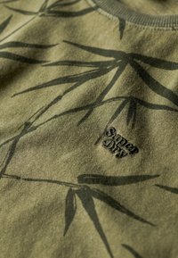 Camiseta de algodón verde con un diseño de hojas de bambú en verde oscuro, con un pequeño logo bordado de "Super Dry" en negro.