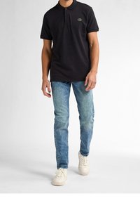 Svart pikétröja med krage, två knappar och en liten logotyp, kombinerad med ljusblå slim-fit denimjeans och vita sneakers.
