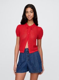 Rød strikket cardigan med korte pufferm, med sløyfedetaljer, knappeåpning og bølgede kanter. Kombinert med høytlivs denimshorts.