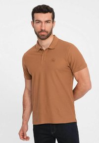 Ruskea polo-paita, jossa on tekstuuri pintakuvio, kaulus ja lyhyet hihat. Hienovarainen logoyksityiskohta rinnassa. Yhdistetty tummiin farkkuihin.