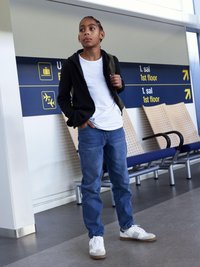 Jeune portant un hoodie noir zippé sur un T-shirt blanc, un jean bleu et des baskets blanches. Un sac à dos vert est porté sur une épaule.