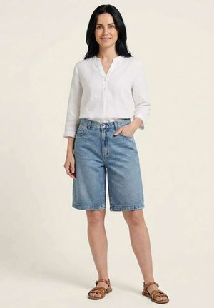 Kvinde iført hvid langærmet skjorte, korte cowboyshorts til knæet og brune sandaler, står med den ene hånd i lommen på ensfarvet baggrund.