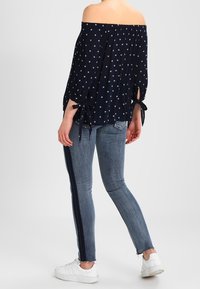 Top navy off-the-shoulder con pois bianchi, scollatura arricciata e dettagli con fiocchi sulle maniche, abbinato a jeans skinny blu con striscia laterale.