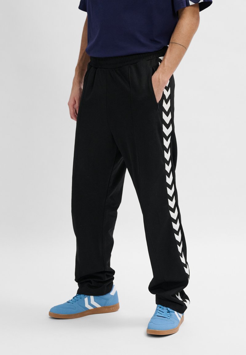 Zwarte trainingsbroek met een elastische tailleband, zijzakken en witte chevronstreepjes langs de zijnaden. Gemaakt in combinatie met blauwe sneakers.