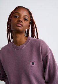 Young woman with long red braids wearing a mauve Carhartt sweatshirt, looking forward against a plain light background.