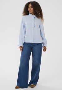 Hellblau gestreiftes Hemd mit geraffter Vorderseite, Knopfverschluss und langen Ärmeln, kombiniert mit weit geschnittenen blauen Jeans und braunen Schuhen.