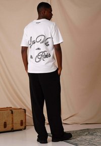 T-shirt bianco in cotone con "La Vie en Rose" stampato in corsivo nero sul retro, abbinato a pantaloni neri larghi e scarpe nere.