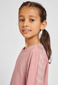 Rosa Langarmshirt mit rundem Ausschnitt, versehen mit einem silbernen Logo und einem Chevron-Muster entlang der Schultern in einem strukturierten Stoff.