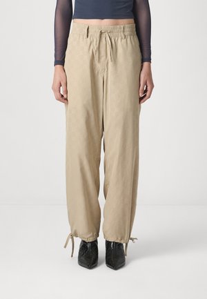 Beige bukser med elastisk talje, snor og teksturert mønster; innsnevrede cuffs med slips. Kombinert med svarte spisse støvler.