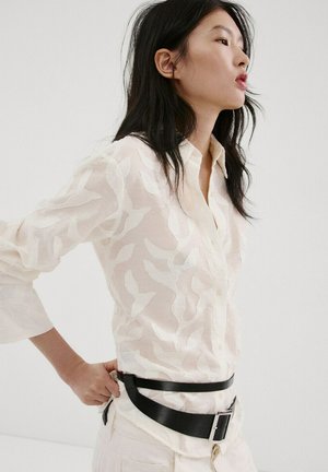 Mujer con el pelo largo y negro, vestida con una camisa blanca transparente con estampado y ajustándose un ancho cinturón de cuero negro alrededor de la cintura.