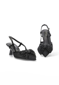 Zapatos de tacón tipo slingback en negro con una parte superior texturizada, que presentan un lazo decorativo de cuentas en la punta y un tacón bajo y cuadrado.