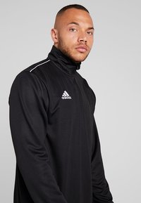 Svart sporty jakke med høy krage, glidelås foran, hvite kontraststriper og Adidas-logo på brystet. Laget av glatt, lett materiale.