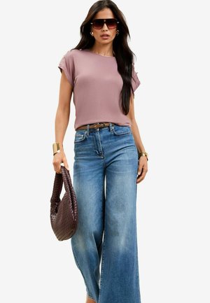Top corto a maniche corte color malva chiaro con risvolti, abbinato a jeans denim blu a gamba larga. Accessoriato con una cintura marrone e una borsa intrecciata scura.