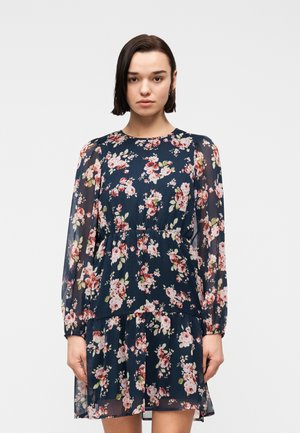 Ung kvinne med kort mørkt hår iført en marineblå blomstrete kjole med lange, gjennomsiktige ermer, stående mot en ensfarget lys bakgrunn.