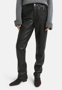 Schwarze Lederhosen mit glatter Oberfläche, ausgestattet mit einem klassischen Fünf-Taschen-Design und einem geraden Bein, kombiniert mit dunklen Stiefeletten.