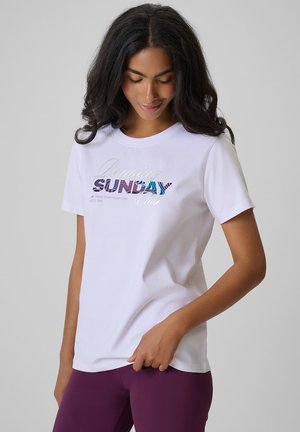 Kobieta o długich czarnych włosach, ubrana w białą koszulkę z wielobarwnym napisem „SUNDAY” i fioletowe legginsy na jednolitym tle.
