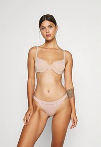 Světlé béžové spodní prádlo, které zahrnuje hladkou podprsenku s nastavitelnými ramínky a ladící nízký tangový kalhotky. Jemná textura s bezešvým provedením.