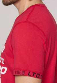 Červené tričko s krátkým rukávem vyrobené z hladké bavlny. Obsahuje černý text na lemu a zpevněné švy podél ramen.