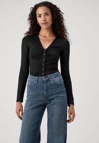 Mulher com cabelo encaracolado vestindo uma blusa preta justa com pregas e botões, de manga longa, e calças jeans azuis de perna larga de cintura alta, em pé contra um fundo branco.