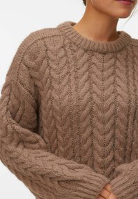 Pull en maille torsadée marron épais porté par une personne les mains jointes devant, montrant la texture et le motif détaillés.