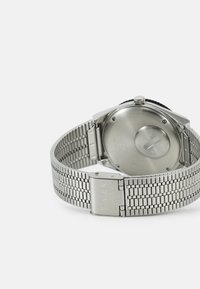 Sølv metalarmbåndsur med meshbånd, der har et rundt urkasse og en fremtrædende krone. Bagsiden viser branding og en glat overflade.