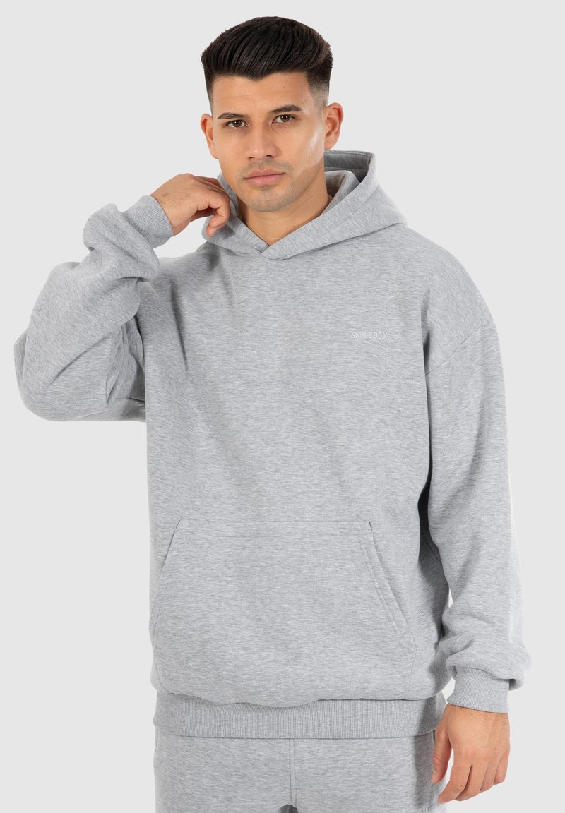 Sweat à capuche gris avec une poche avant, des poignets et un ourlet côtelés. Présente un petit logo sur la poitrine. Le tissu semble doux et texturé.